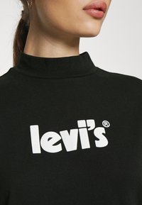 Gros plan d'une personne portant un haut à col montant noir avec un logo Levi's blanc sur la poitrine, montrant les lèvres et une partie de la mâchoire.