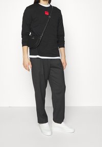 Mann trägt ein schwarzes Langarmshirt mit rotem Logo, dunkelgraue Hose, weiße Sneakers und eine schwarze Umhängetasche vor einem weißen Hintergrund.