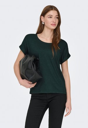 Frau mit langen braunen Haaren, die ein dunkelgrünes Kurzarmoberteil und schwarze Hose trägt und eine große schwarze Lederhandtasche unter dem Arm hält.