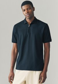 Tricou polo negru cu guler cu fermoar, având un model cu diamante texturat. Mâneci scurte și un design ajustat.