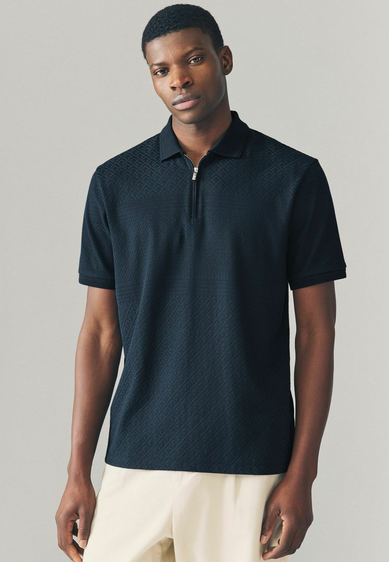 Tricou polo negru cu guler cu fermoar, având un model cu diamante texturat. Mâneci scurte și un design ajustat.