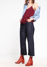 Denim weitem Bein Hosen, bordeauxfarbene Spaghettiträgerbluse, leichte Jeansjacke und rote Absatzstiefeletten. Glatte Texturen, moderne Passform und lebendige Farben.