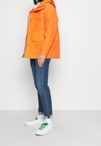 Chaqueta impermeable de color naranja brillante con capucha, múltiples bolsillos y puños ajustables, combinada con jeans azules y zapatos blancos con cordones verdes.