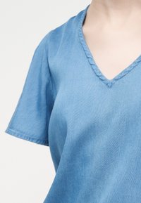 Κοντινό πλάνο ενός ατόμου που φοράει ένα ξεθωριασμένο ανοιχτό μπλε ντένιμ τοπ με V-λαιμόκοψη και κοντά μανίκια, πάνω σε απλό λευκό φόντο.