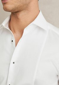 Cămașă albă dintr-un material texturat, cu guler deschis, butoane negre vizibile și un design adaptat, care lasă parțial la vedere gulerul și gâtul.