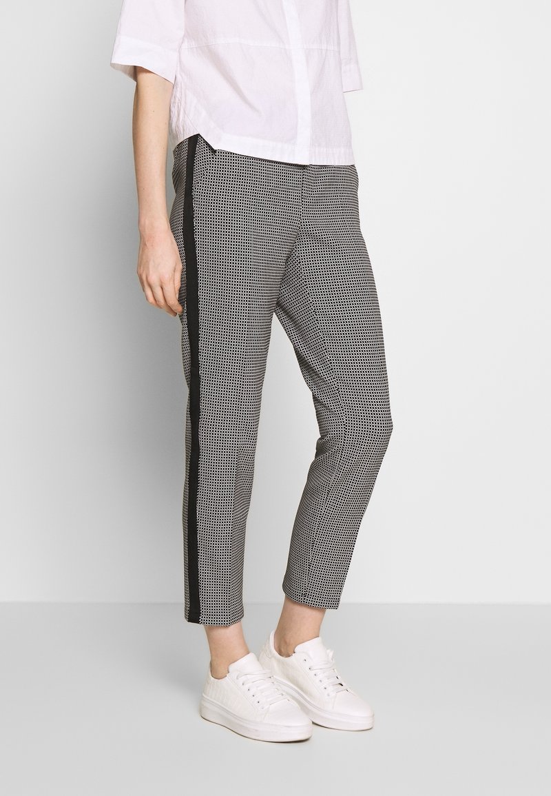 Personne portant un pantalon slim à carreaux noir et blanc avec des bandes latérales noires, des baskets blanches et une chemise blanche à manches courtes sur un fond uni.