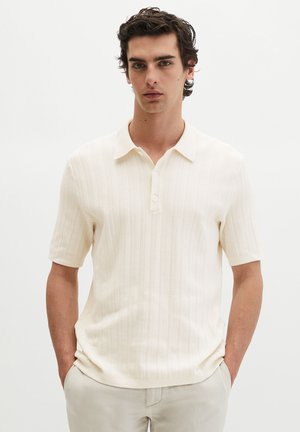 Młody mężczyzna o ciemnych, kręconych włosach, ubrany w kremową, krótkim rękawem dzianinową koszulkę polo i jasne spodnie, ręce w kieszeniach.