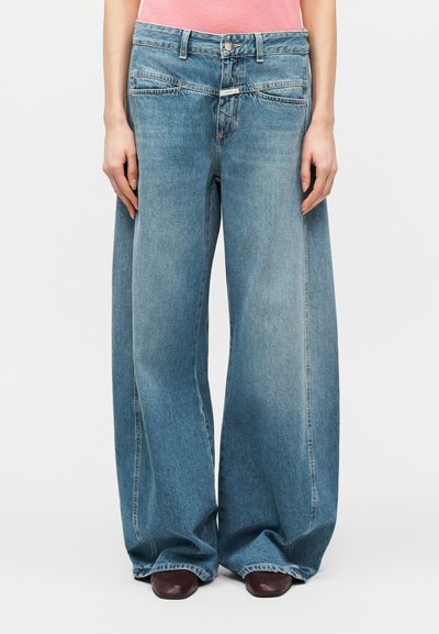Personne portant un jean large bleu à évasement exagéré qui couvre presque des chaussures noires à bout carré, associée à un haut rose.