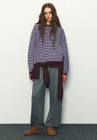 Pruhovaný sveter v fialovej a svetlomodrej farbe, skombinovaný s voľnými sivými nohavicami. Bordová košeľa zviazaná v páse a hnedé mokasíny.