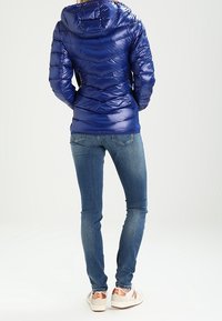 Blå pufferjakke med hætte, prydet med et chevron-design og glansfuld overflade. Bæret med tætsiddende jeans og hvide sneakers.