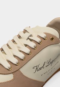 Κοντινό πλάνο μπεζ και κρεμ αθλητικού παπουτσιού με λευκά κορδόνια και χρυσά περφορετ, με το λογότυπο «Karl Lagerfeld» και τη διεύθυνση του Παρισιού στο πλάι.