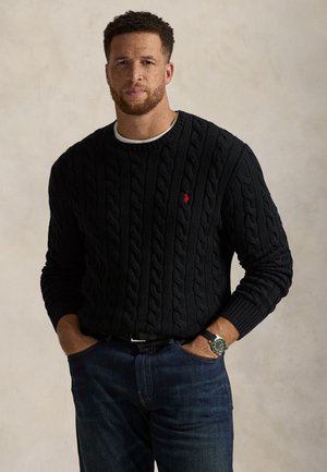 Homme aux cheveux courts et bouclés avec une barbe, portant un pull en maille torsadée noire avec un petit logo rouge et un jean bleu, mains dans les poches.
