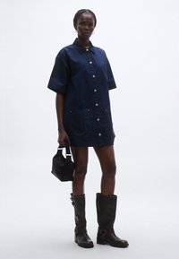 Tengerészkék, rövid ujjú ingruha gombos záródással és első zsebekkel; magas fekete bőr csizmával és kis kézitáskával párosítva.