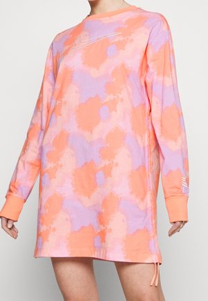 Sukienka z długim rękawem w stylu tie-dye w pastelowym różu, pomarańczy i purpurze z małym logo Nike na piersi i nadgarstku, noszona przez stojącą osobę.