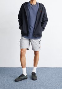 Mężczyzna w czarnej kurtce na zamek, noszący niebieską koszulę, jasnoszare spodenki cargo, białe skarpetki i ciemne sneakersy, stojący na niebieskim dywanie.