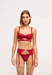 Czerwony koronkowy komplet bielizny, zawierający biustonosz z fiszbinami i złotymi akcentami, pasujące majtki oraz pas do pończoch z paskami. Przezroczyste cieliste pończochy do uda.