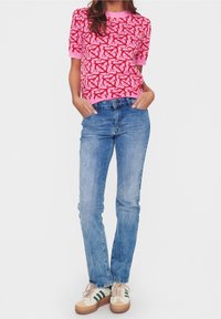 Pink short-sleeve sweater with red abstract pattern, paired with blue denim jeans and beige sneakers featuring green stripes.