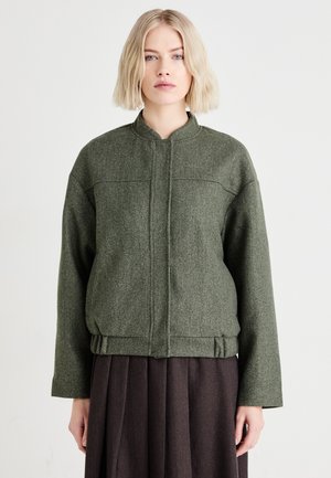 Veste en laine verte à col mandarin, épaules tombantes et taille cintrée. Texture lisse avec des détails de couture minimaux.