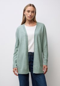 Junge Frau, die eine hellgrüne, offene Strickjacke über einem weißen Oberteil und dunkelblauen Jeans trägt, vor einem hellen Hintergrund.