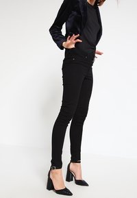 Černý zkrácený sametový blazer přes černý přiléhavý top a černé skinny džíny. Modelka má na sobě lesklé černé lodičky s hladkým povrchem.