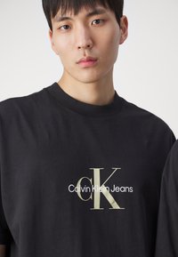 Černé bavlněné tričko s logem Calvin Klein Jeans ve zlaté a bílé barvě. Kulatý výstřih a krátké rukávy. Hladká textura s volným střihem.