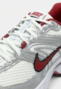 Zapatilla de running Nike con parte superior de malla plateada y detalles en rojo, cordones blancos y un prominente logo Swoosh en negro y rojo. Diseño ligero.