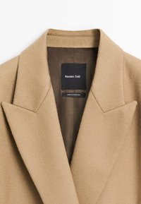 Camel-colored blazer with a structured design, made of textured fabric. Features sharp lapels and a black label with brand name inside.