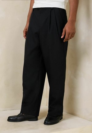 Mann, der lose schwarze plissierte Hosen und schwarze Lederloafers trägt, steht vor einer beigen Wand und einem beigen Boden.