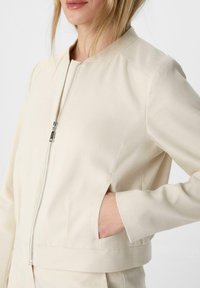 Beżowa kurtka zapinana na zamek z okrągłym dekoltem, krótkim krojem i bocznymi kieszeniami. Wykonana z teksturowanej tkaniny o gładkim wykończeniu.