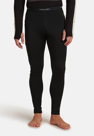 Personne portant des leggings Icebreaker noirs ajustés et un haut à manches longues noir avec des manches beiges, se tenant pieds nus sur un fond blanc.