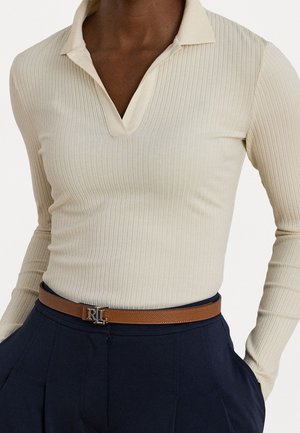 Personne portant un polo à manches longues côtelé crème, rentré dans un pantalon bleu marine avec une ceinture en cuir marron avec une boucle métallique RL.