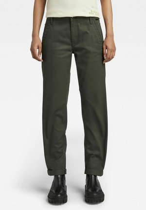 Personne portant un pantalon vert foncé à jambes droites roulé aux ourlets avec des bottines noires épaisses et une chemise beige clair.