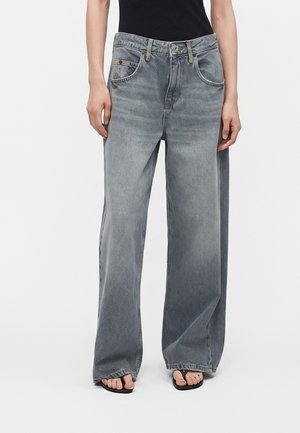 Person iført brede, falmede grå jeans med en sort top og sorte sandaler, står mod en ensfarvet hvid baggrund.