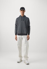 Tmavý sweatshird s vzorom a šedým motívom blesku, nosený s bielymi voľnými nohavicami a šedými teniskami. Čiapka dodáva akcent k neformálnemu oblečeniu.