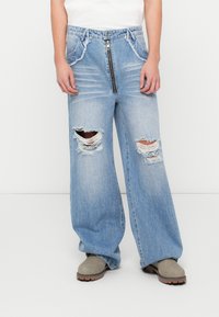 Laialised helesinised denim püksid, millel on ees tõmblukk, kahjustatud detailid suurte rebenditega ja kerge pleekimine. Kanda hallide pahkluupükstega saapadega.