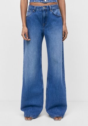 Blå vidde jeans båret af en person med crop-top i denim og firkantede sandaletter, stående mod en ensfarvet hvid baggrund.