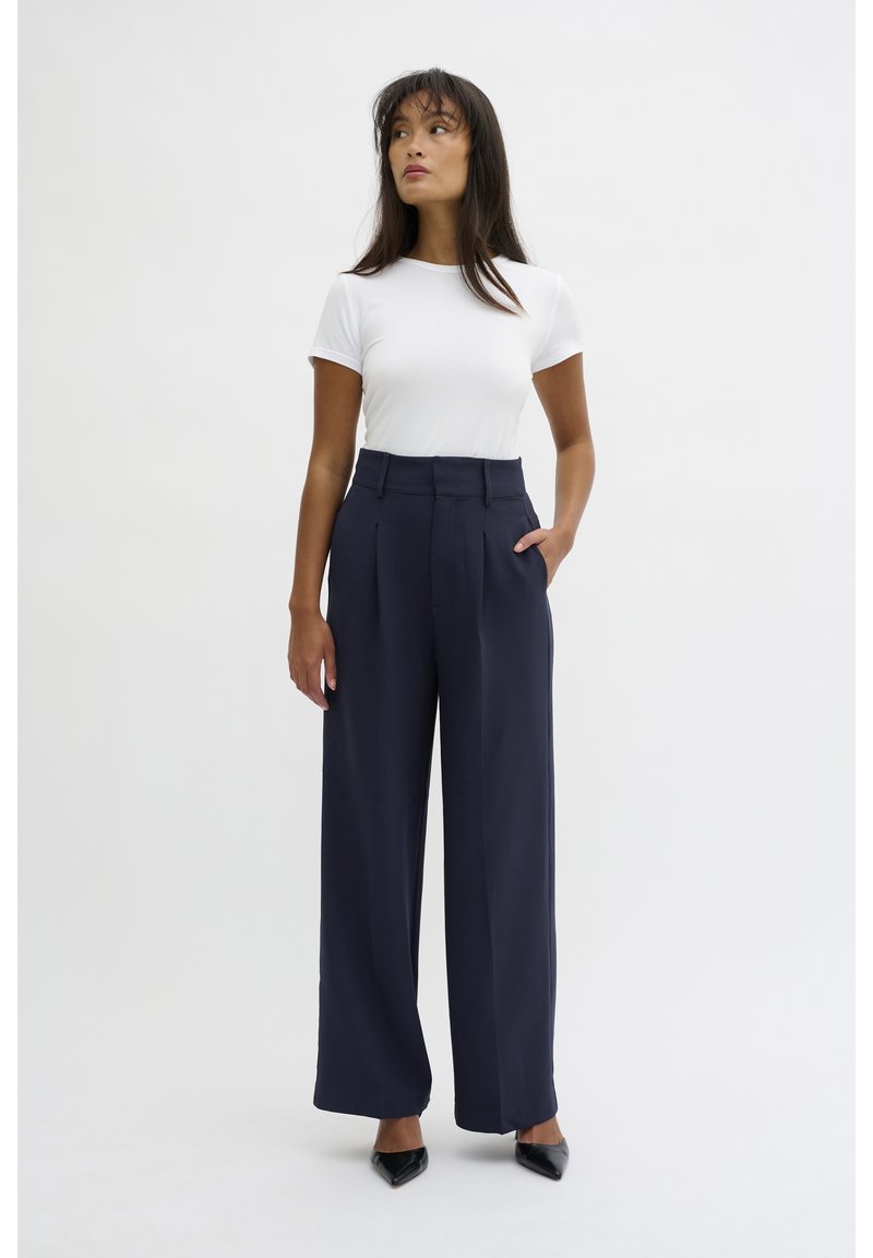 High-waisted navy trousers with pleats, wide legs, and a smooth texture paired with a fitted white t-shirt and black pointed heels.