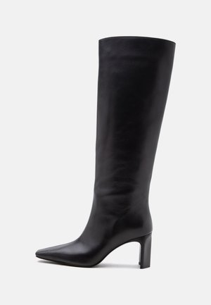 Botte en cuir noire montant jusqu'au genou avec bout pointu et talon bloc sur fond clair.