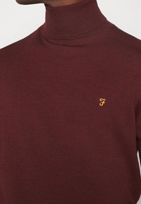 Weinroter Rollkragenpullover aus geripptem Strickstoff. Verfügt über ein kleines gelbes Logo auf der Brust, einen eng anliegenden Kragen und eine weiche Textur.