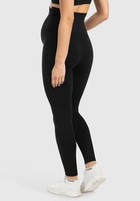 Leggings noirs taille haute en tissu extensible, avec une texture lisse et un design ajusté, accompagnés de chaussures de sport blanches.