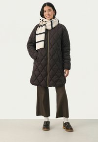 Quiltad brun jacka med gulddragkedja, svart-vit randig scarf, bruna vida byxor och svarta skor med tjock sula.