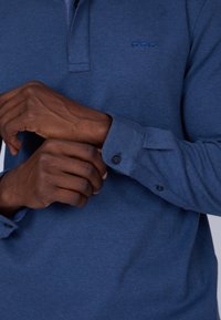 Polo à manches longues bleu marine en tissu texturé. Doté d'une patte de boutonnage à deux boutons et de poignets boutonnés. Petit logo brodé sur la poitrine.