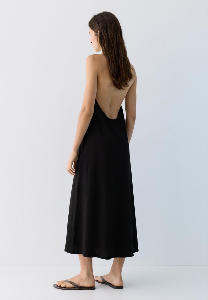 Femme debout, dos visible, portant une robe midi noire sans manches et dos nu, avec des sandales grises sur un fond clair uni.