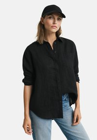 Zwarte linnen button-up shirt met een relaxte pasvorm, opgerolde mouwen en een subtiele pocketdetail, gecombineerd met een blauwe denim jeans en een zwarte pet.