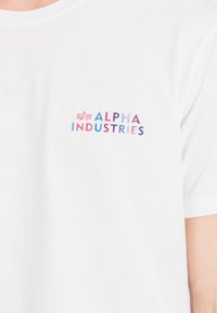 Camiseta blanca de algodón con cuello redondo que presenta un logo multicolor de "ALPHA INDUSTRIES" en la parte izquierda del pecho. Textura suave y corte casual.