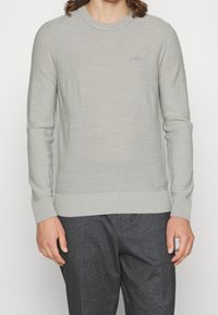 Man som bär en ljusgrå tröja med rund hals och "BOSS"-logotyp, samt mörkgrå byxor med prickar, avkortade vid halsen och mitt på låret.