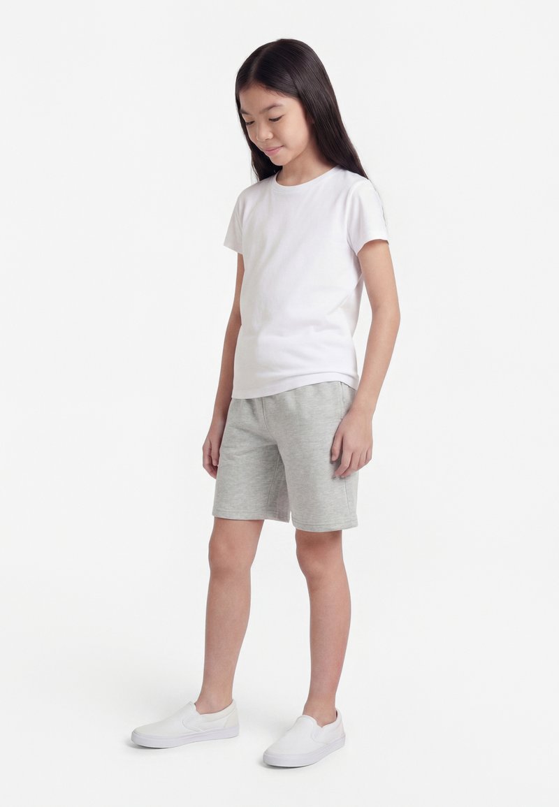 Niña joven de pie, vestida con una camiseta blanca lisa, pantalones cortos grises hasta la rodilla y zapatos blancos sin cordones sobre un fondo blanco.