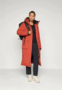 Lungo cappotto rosso con cappuccio, caratterizzato da numerose tasche, accenti neri e un design aerodinamico. Indossato su un outfit nero, abbinato a scarpe da ginnastica.