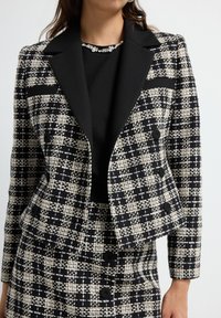 Blazer a scacchi bianco e nero con colletto scialle nero, doppio bottone e tessuto testurizzato, abbinato a una maglietta nera senza maniche sotto.