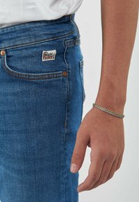 Jeans di denim blu con una vista ravvicinata di una mano che indossa un bracciale in strass argentato. Presentano un'etichetta di marca sulla tasca posteriore.